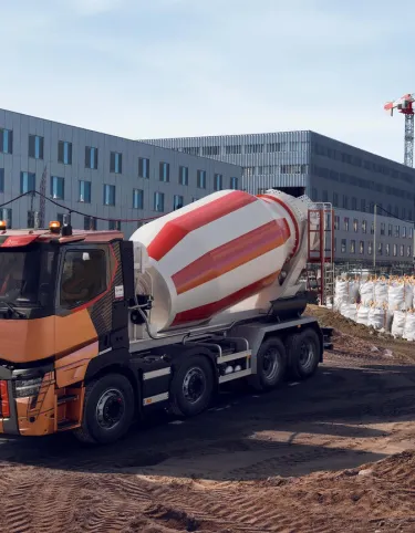 A Renault Trucks C lorry fitted with a concrete mixer is operating on a large urban construction site surrounded by cranes and modern buildings.