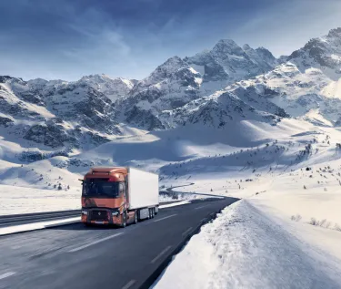 A Renault Trucks T High tractor unit pulling a refrigerated semi-trailer is travelling along a winding mountain road amidst a majestic snow-covered landscape.